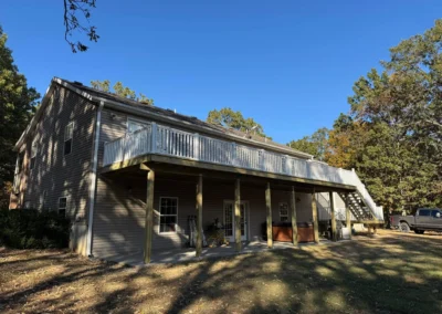 A two-story house with a wooden deck, surrounded by trees and a clear blue sky, featuring a truck parked nearby.